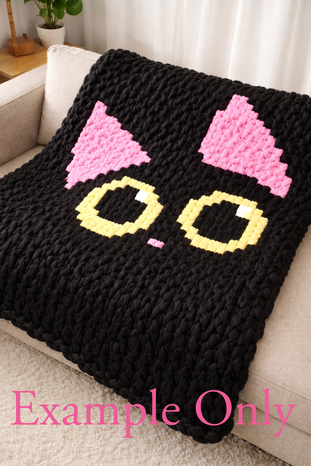 Black knitted blanket with pink cat ears and yellow circle eyes on a beige couch.