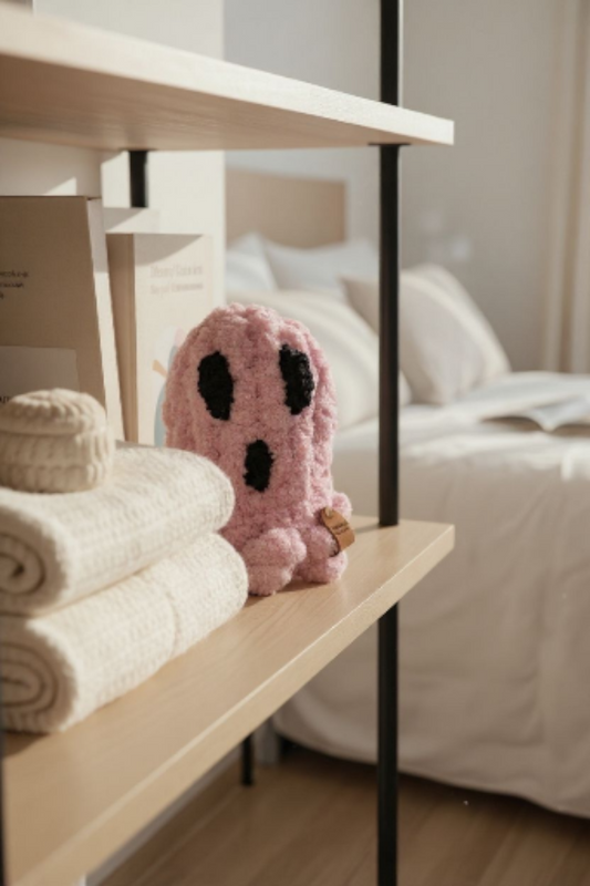 Pink ghost-shaped pillow on a shelf with folded towels, bed in the background
