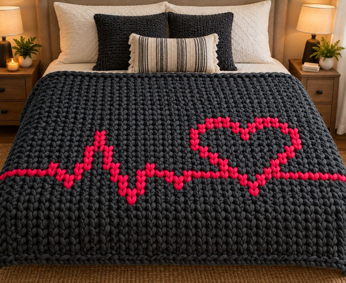 A chunky knit blanket in dark charcoal gray with a bright pink heartbeat line and heart design, styled on a bed with white and striped pillows in a warmly lit bedroom.