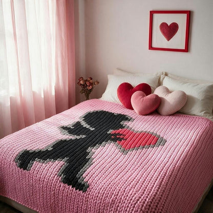 A pink knit blanket with a large black and red heart design is spread across a bed in a softly lit bedroom. White pillows and three heart-shaped throw pillows rest on the bed, with a pink heart framed print on the wall above.