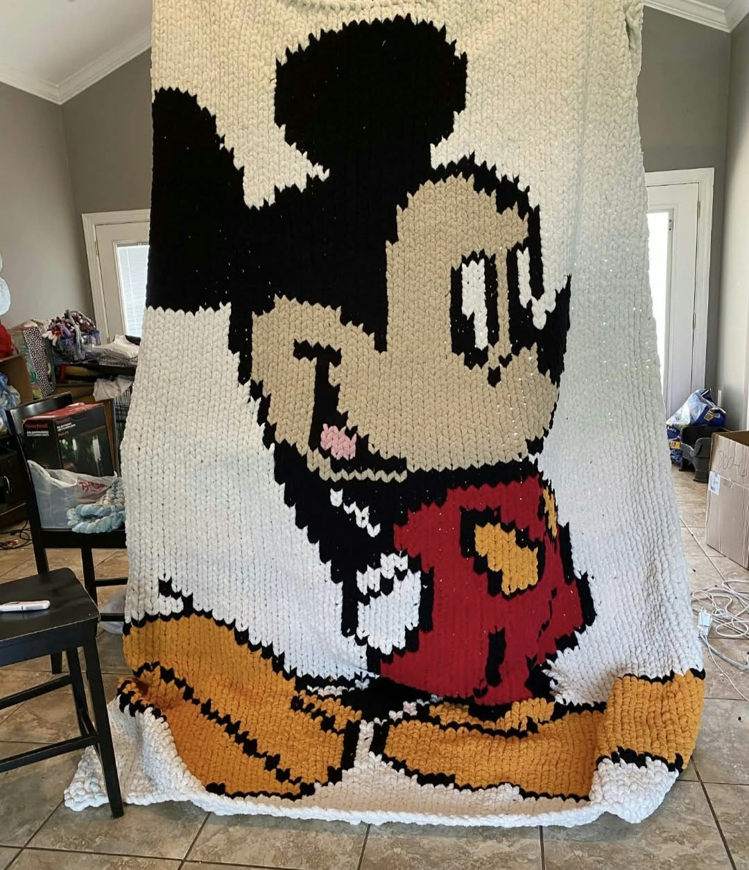 A large hand-knit blanket featuring a pixelated Mickey Mouse design, hanging in a home interior room.
