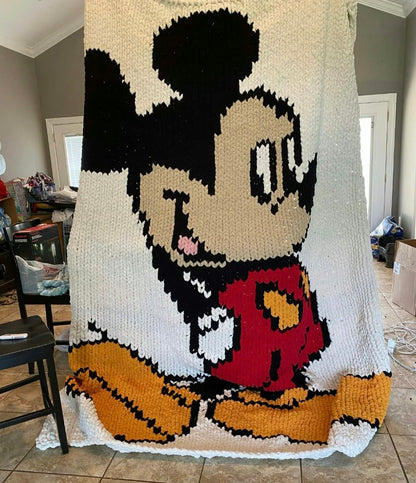 A large hand-knit blanket featuring a pixelated Mickey Mouse design, hanging in a home interior room.
