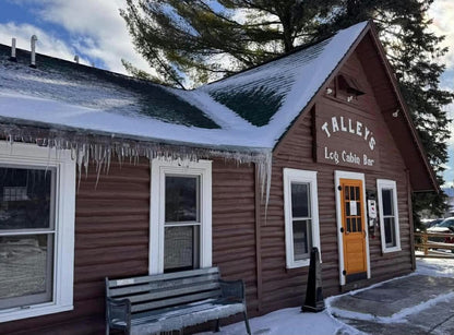 A rustic wooden cabin bar with snow on the roof and icicles hanging from the eaves, featuring a sign that reads 'TALLEY'S LCG. CABIN BAR' above an orange door.