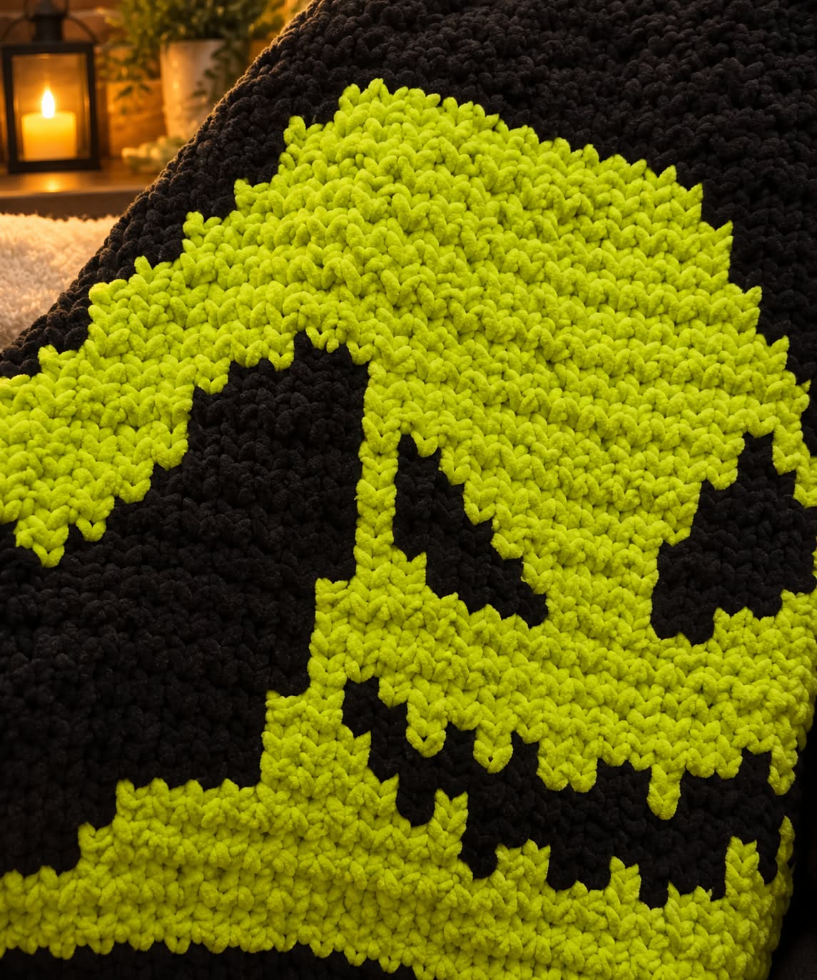 Close-up of a chunky knit blanket featuring a bright Oogie Boogie face design on a black background, with a softly blurred warm indoor scene in the background.