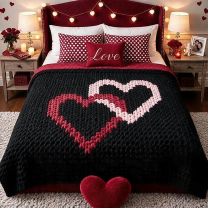 A cozy bedroom scene featuring a black chunky knit blanket with a large knitted heart design in red and white, styled with matching red and white polka dot pillows and a red 'Love' accent pillow on a dark red headboard.