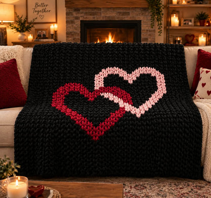 A chunky knit throw blanket draped over a white sofa in a cozy living room. The blanket is black with two large interlocking hearts in red and light pink.