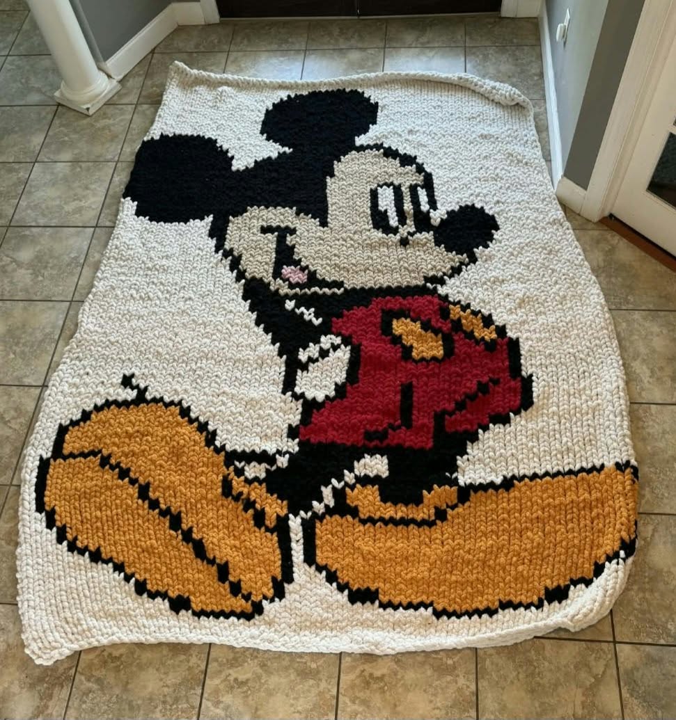 A handmade, chunky knitted Mickey Mouse rug laid out on a tiled floor. The design features a pixelated Mickey Mouse in his classic red shorts, yellow shoes, and black ears, set against a cream-colored knitted background.