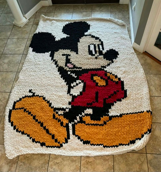 A handmade, chunky knitted Mickey Mouse rug laid out on a tiled floor. The design features a pixelated Mickey Mouse in his classic red shorts, yellow shoes, and black ears, set against a cream-colored knitted background.