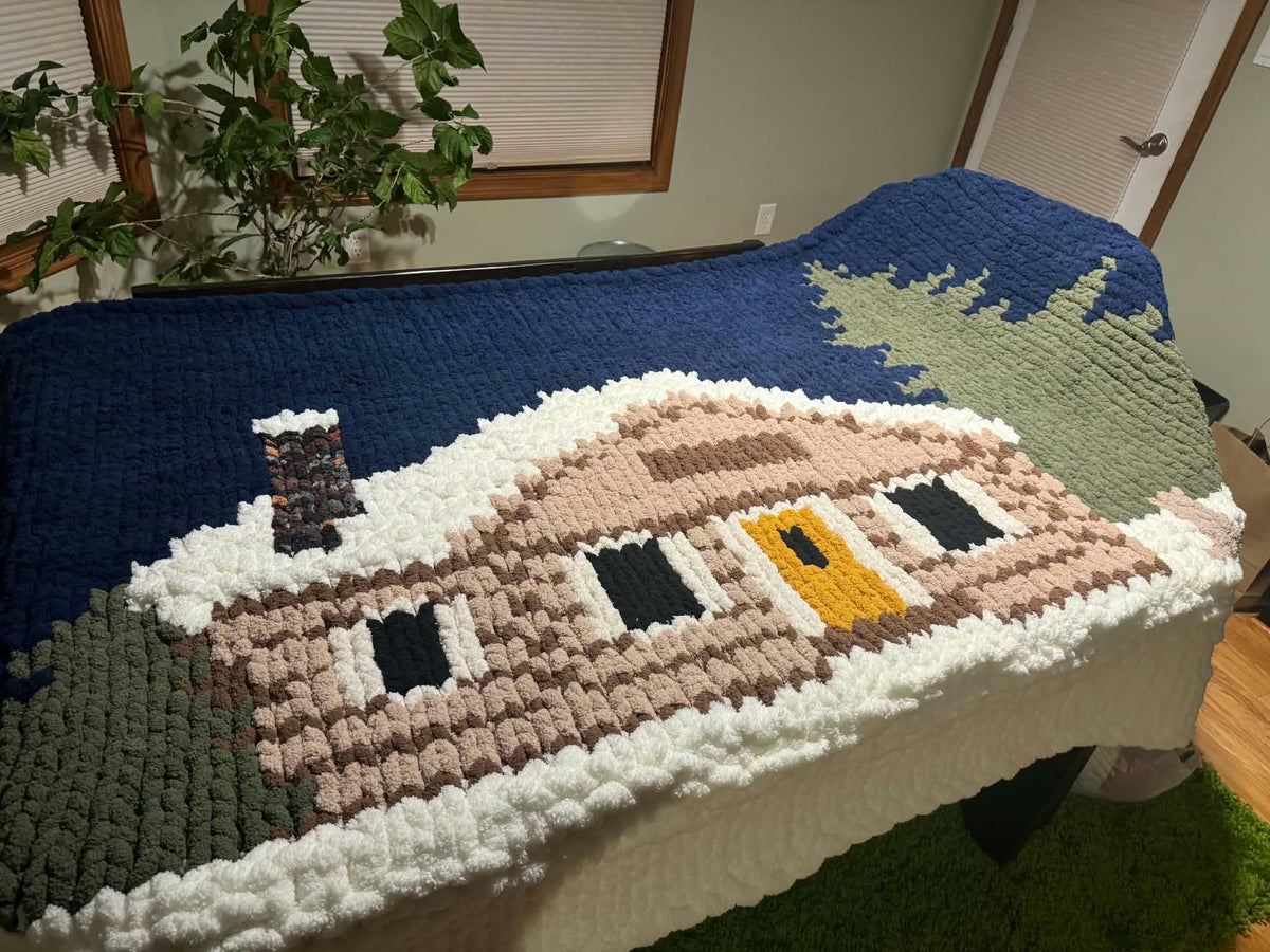 A large chunky knit blanket draped over a table in a home setting, featuring a detailed log cabin design with a blue sky, green trees, and a yellow front door.