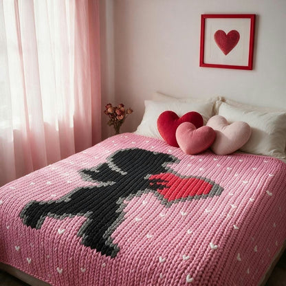 A cozy bedroom scene with a pink knitted blanket featuring a black silhouette of a person holding a red heart, styled on a bed with pink and red heart-shaped pillows and a framed heart print on the wall.