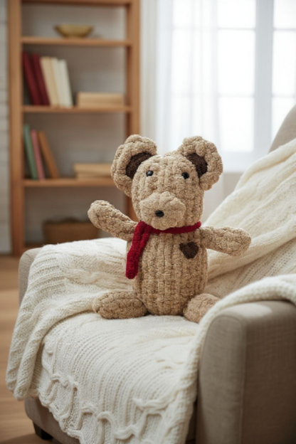 Knitted teddy bear with a red scarf sitting on a textured blanket in a cozy room.