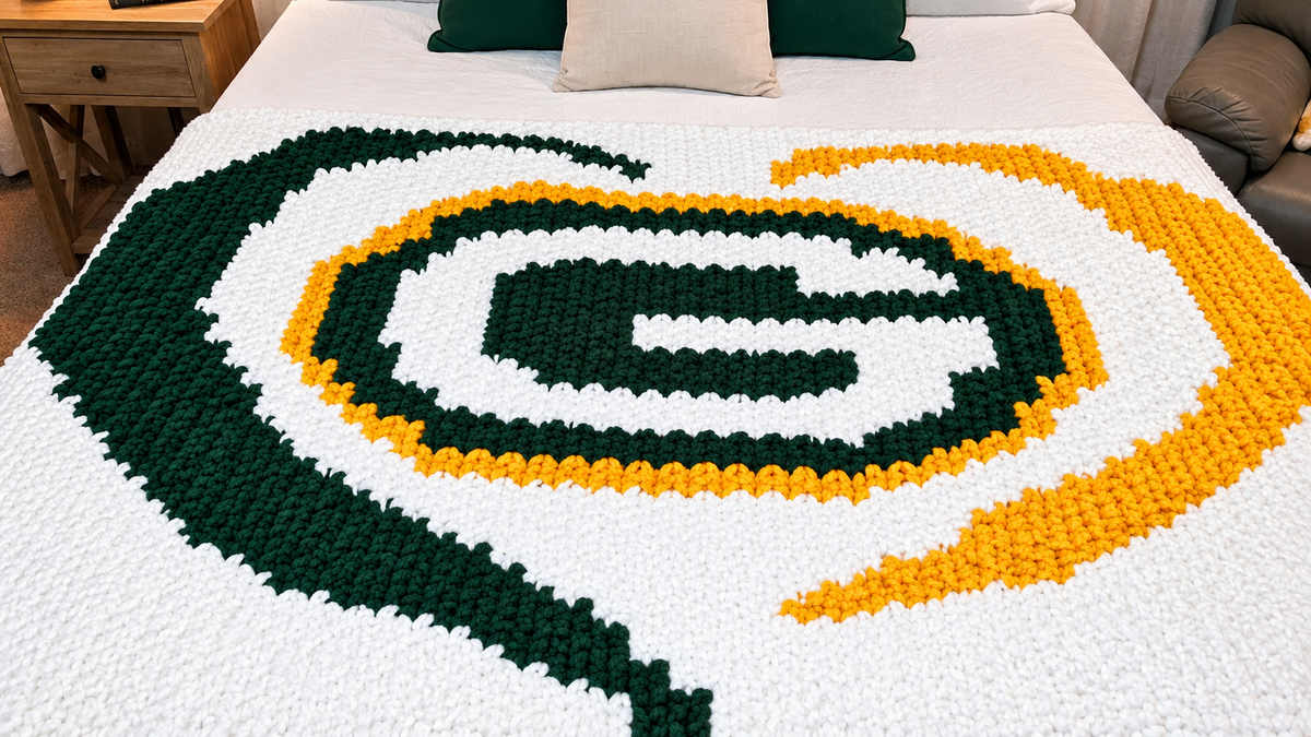 A large, chunky knit blanket,  Green Bay Packers-themed blanket spread across a bed, featuring a white base with a green and yellow logo pattern. The bed is in a bedroom setting with pillows and a bedside table visible.