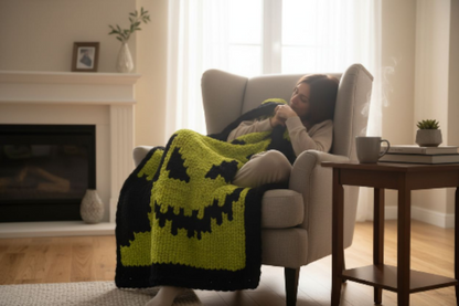 Person sitting in a chair with a green and black patterned blanket, in a cozy living room setting.